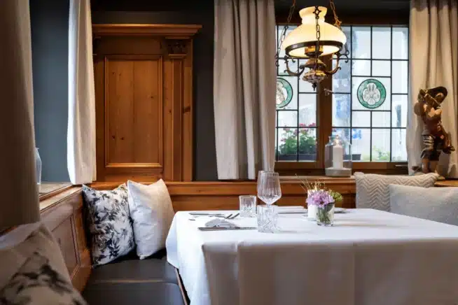 Eine gemütliche Restaurant-Essecke mit Holzvertäfelung, weißer Tischdecke, gedecktem Geschirr, dekorativen Kissen und Blumen in einer Vase. Von oben hängt warmes Licht herab, und große Fenster lassen sanftes Tageslicht herein.