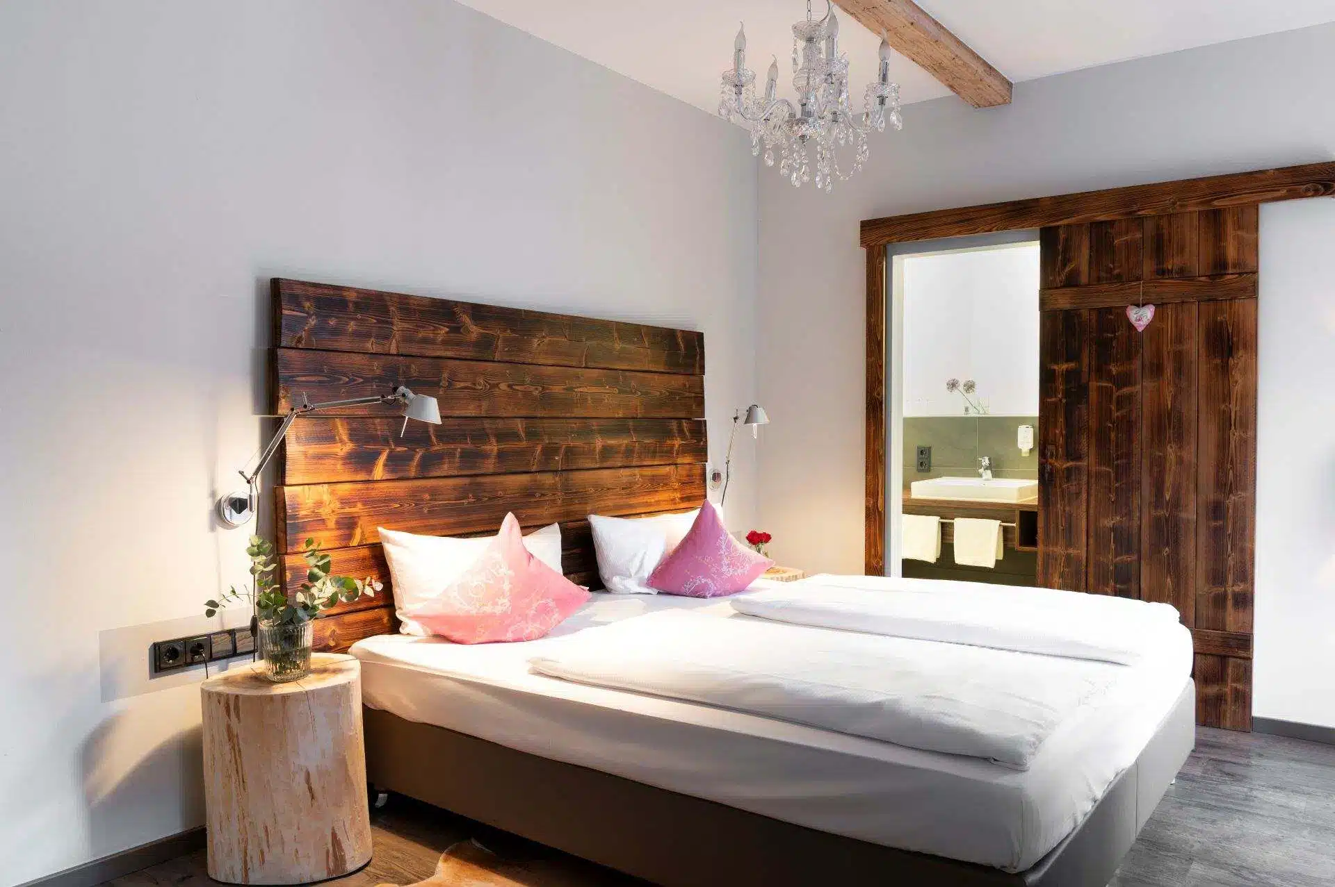 Ein gemütliches Schlafzimmer mit einem Doppelbett, einem hölzernen Kopfteil, zwei rosafarbenen Akzent-Kissen und modernen Lampen. Ein Baumstumpf dient als Nachttisch. Durch eine offene Tür gelangt man in ein Badezimmer mit Waschbecken und Blumen.