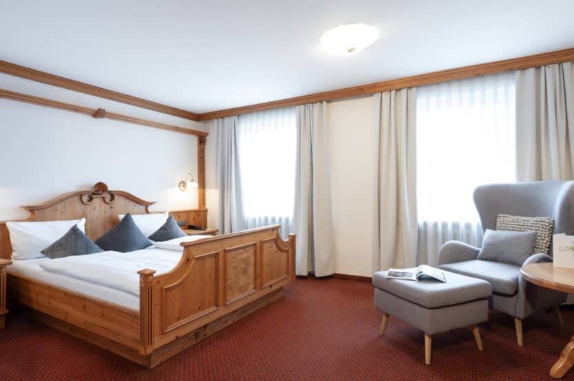 Ein gemütliches Hotelzimmer mit einem hölzernen Doppelbett, grauen Akzentkissen, zwei großen Fenstern mit hellen Vorhängen, einem grauen Sessel mit Ottomane und einem kleinen Tisch mit einem aufgeschlagenen Buch und einer Speisekarte darauf.