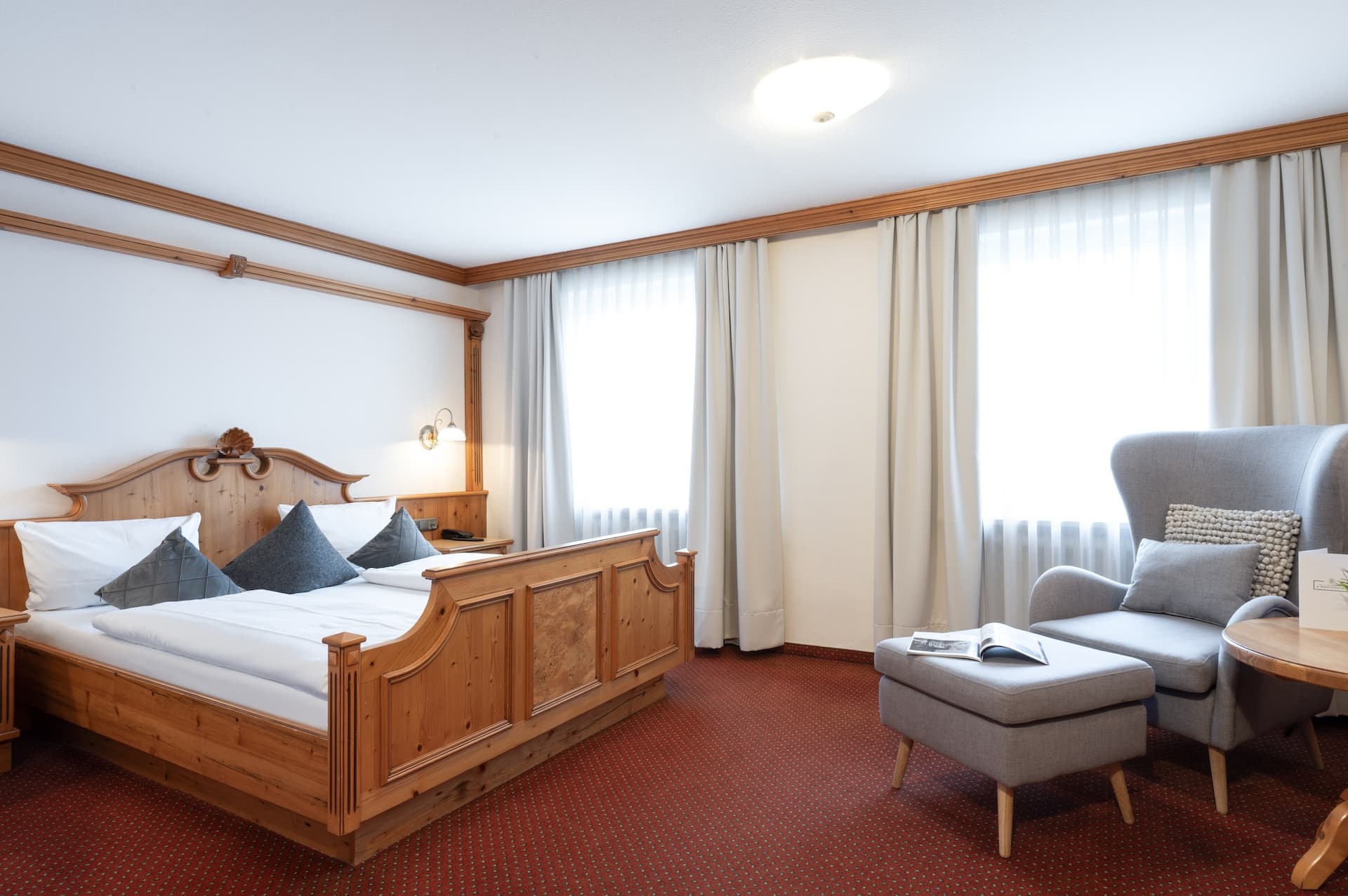 Ein gemütliches Hotelzimmer mit einem hölzernen Doppelbett, grauen Akzentkissen, zwei großen Fenstern mit hellen Vorhängen, einem grauen Sessel mit Ottomane und einem kleinen Tisch mit einem aufgeschlagenen Buch und einer Speisekarte darauf.