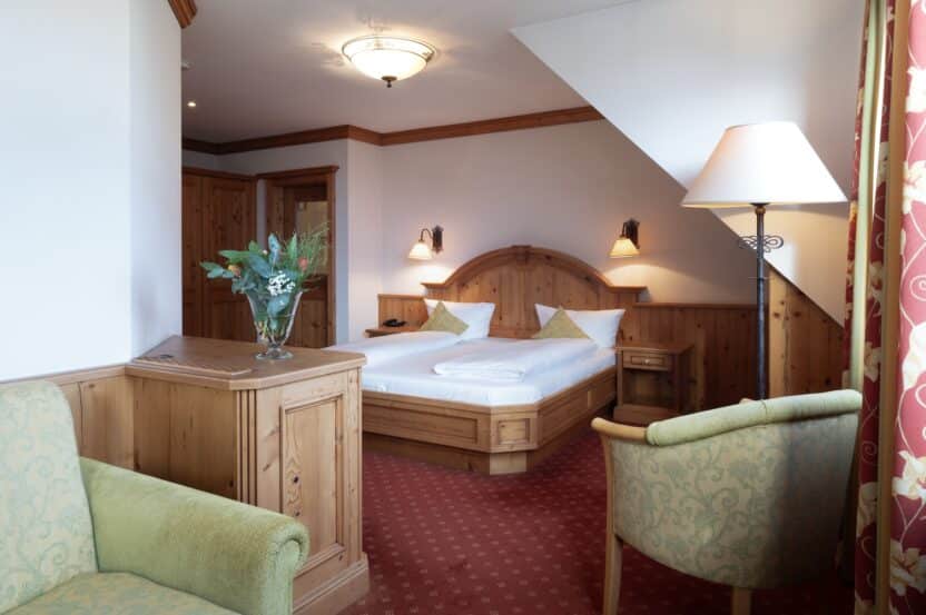 Ein gemütliches Hotelzimmer mit einem Doppelbett mit weißer Bettwäsche, Holzmöbeln, einem grünen Sessel, einer Stehlampe und einer Vase mit Blumen auf einem Holztisch. Das Zimmer hat einen roten Teppich und eine warme Beleuchtung.