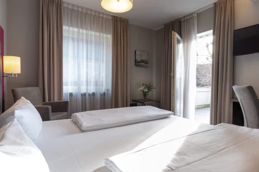 Helles Hotelzimmer mit einem Doppelbett, zwei Stühlen, einem kleinen Tisch mit Blumen, großen Fenstern mit beigen Vorhängen, einem an der Wand montierten Fernseher und einer offenen Tür, die auf einen Balkon führt.