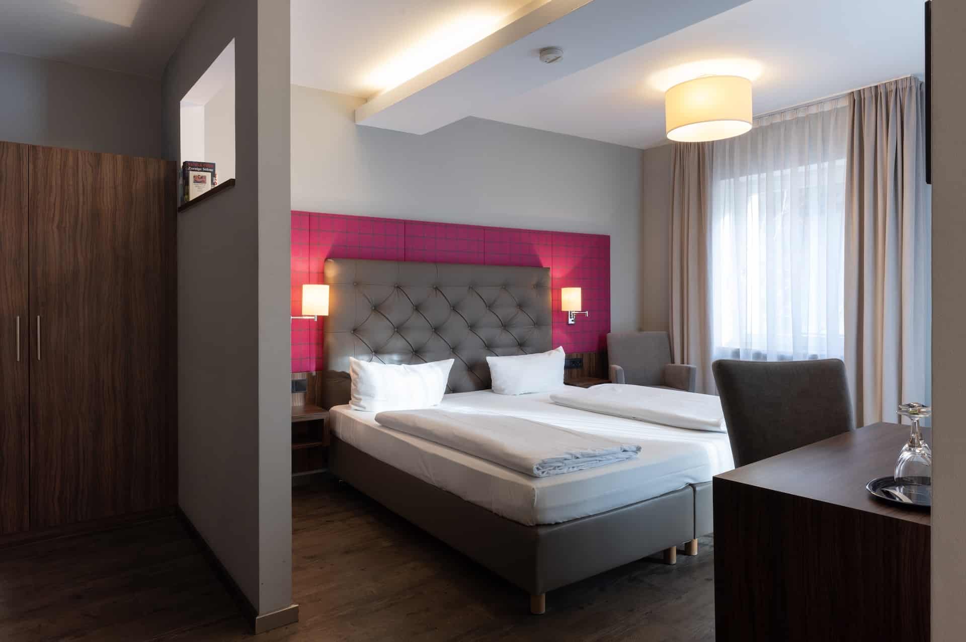 Modernes Hotelzimmer mit einem Doppelbett, getuftetem Kopfteil, zwei Nachttischlampen, Stühlen am Fenster mit transparenten Vorhängen, Holzboden und einem Kleiderschrank. Das Zimmer ist minimalistisch eingerichtet und warm beleuchtet.