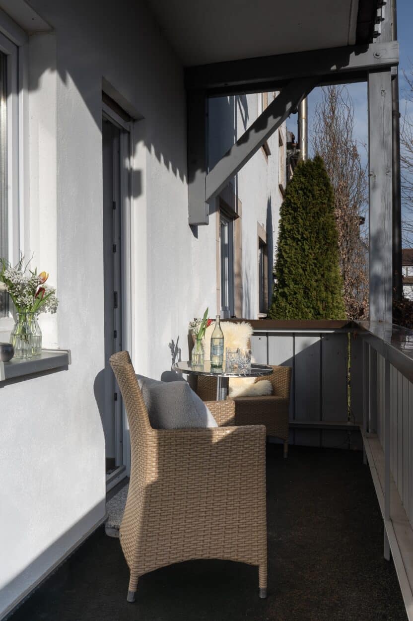 Ein kleiner Balkon mit zwei Korbsesseln, einem Tisch mit Getränken und Blumen, und Sonnenlicht, das Schatten wirft. Es gibt Topfpflanzen, ein Fenster mit einer Blumenvase und einen hohen grünen Busch im Hintergrund.