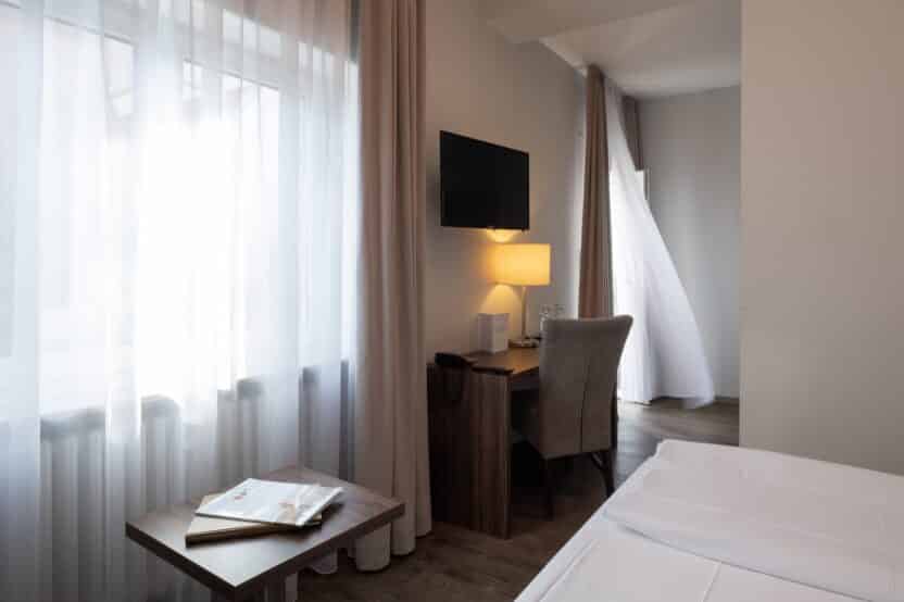 Ein modernes, sonnendurchflutetes Hotelzimmer mit durchsichtigen Vorhängen, einem hölzernen Schreibtisch und Stuhl, einem an der Wand montierten Fernseher, einer Lampe und einem ordentlich gemachten Bett; eine leichte Brise hebt einen Vorhang an der Balkontür.