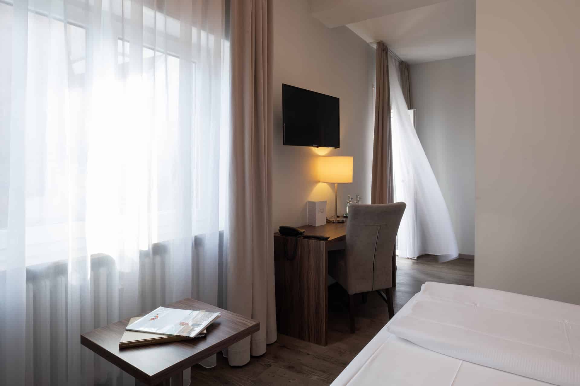 Ein modernes, sonnendurchflutetes Hotelzimmer mit durchsichtigen Vorhängen, einem hölzernen Schreibtisch und Stuhl, einem an der Wand montierten Fernseher, einer Lampe und einem ordentlich gemachten Bett; eine leichte Brise hebt einen Vorhang an der Balkontür.