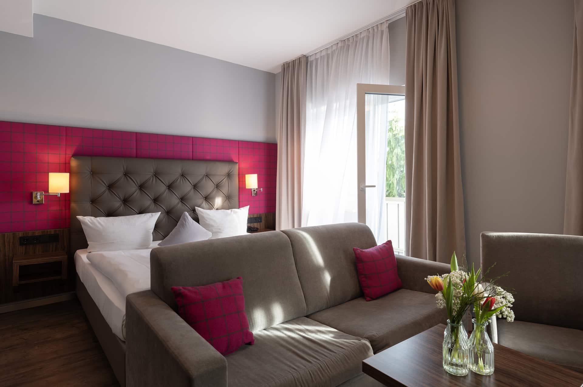 Ein modernes Hotelzimmer mit einem Doppelbett, einem taupefarbenen Kopfteil, einem grauen Sofa, roten Akzentkissen, einem Holztisch mit einer Blumenvase und großen Fenstern mit beigen Vorhängen, die natürliches Licht hereinlassen.