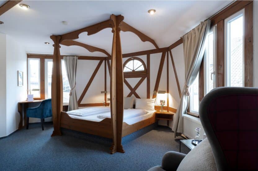 Ein gemütliches Hotelzimmer mit einem Himmelbett aus Holz, zwei Nachttischen mit Lampen, großen Fenstern mit Vorhängen, einem Schreibtisch mit einem blauen Stuhl und einem Sessel im Vordergrund. Der Raum ist von natürlichem Licht durchflutet.