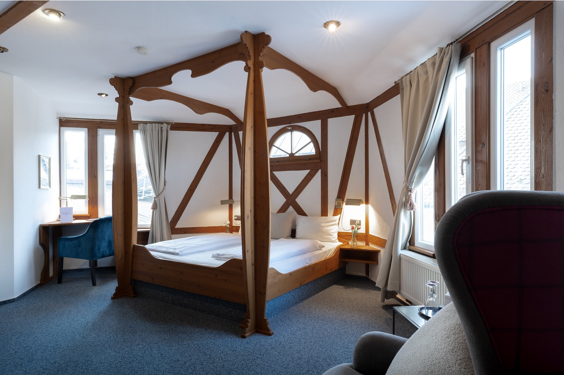 Ein gemütliches Hotelzimmer mit einem Himmelbett aus Holz, zwei Nachttischen mit Lampen, großen Fenstern mit Vorhängen, einem Schreibtisch mit einem blauen Stuhl und einem Sessel im Vordergrund. Der Raum ist von natürlichem Licht durchflutet.