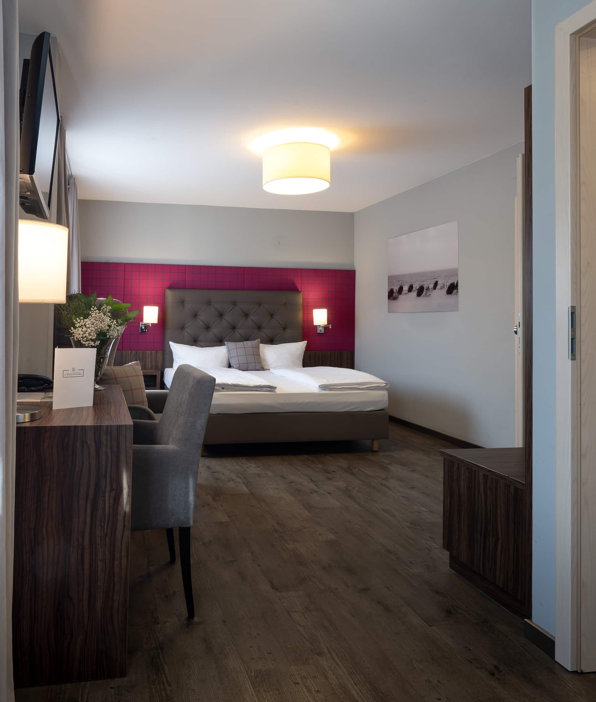 Modernes Hotelzimmer mit großem Bett, grauem Polsterkopfteil, magentafarbener Akzentwand, Schreibtisch mit Stuhl, Blumen auf dem Schreibtisch, an der Wand montiertem Fernseher und sanfter Deckenbeleuchtung; Holzfußboden im gesamten Zimmer.