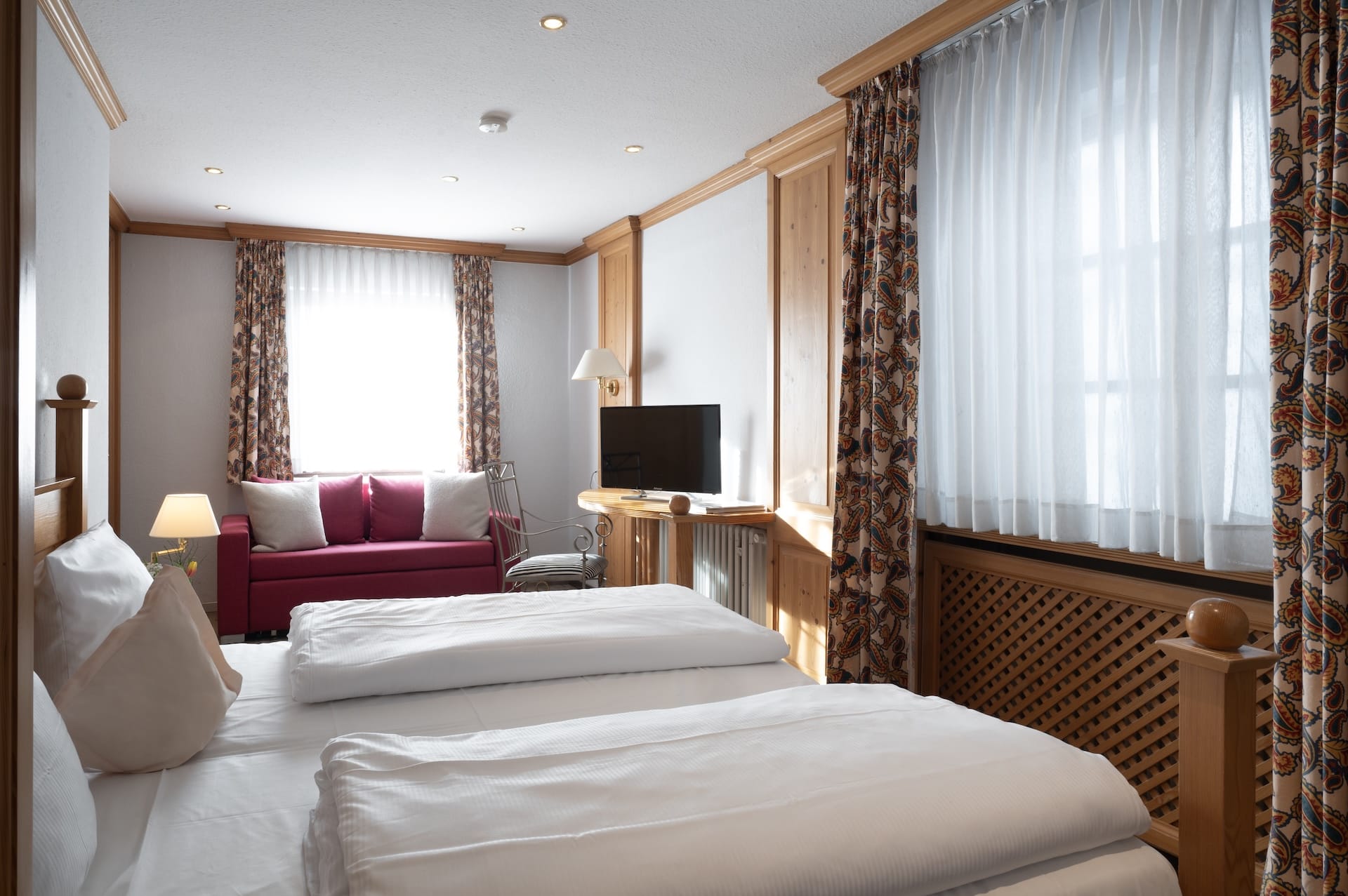 Ein gemütliches Hotelzimmer mit zwei Einzelbetten, weißer Bettwäsche, holzgetäfelten Wänden, geblümten Vorhängen, großen Fenstern, einem kleinen Fernseher auf einem Holzschrank und einem rosafarbenen Sofa unter einem Fenster mit durchsichtigen weißen Vorhängen.