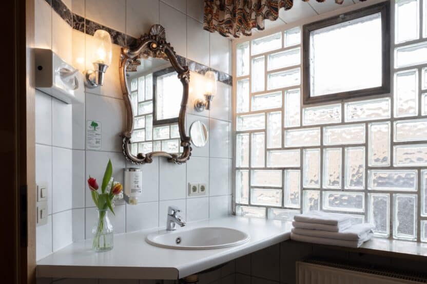 Ein helles Badezimmer mit weißen Fliesenwänden, einem dekorativen Spiegel, Wandleuchten, einer Tulpenvase auf dem Tresen, gestapelten Handtüchern und einem Fenster aus Glasbausteinen, das natürliches Licht hereinlässt.