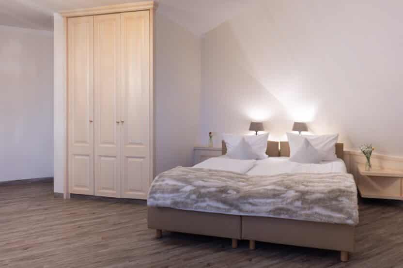 Ein gemütliches Schlafzimmer mit einem großen Bett, das mit einer Pelzdecke bedeckt ist, zwei weißen Kissen, einem hölzernen Kleiderschrank, Nachttischen mit Lampen und Blumen in Vasen. Der Boden ist aus Holz und die Wände sind in einem sanften Beigeton gehalten.