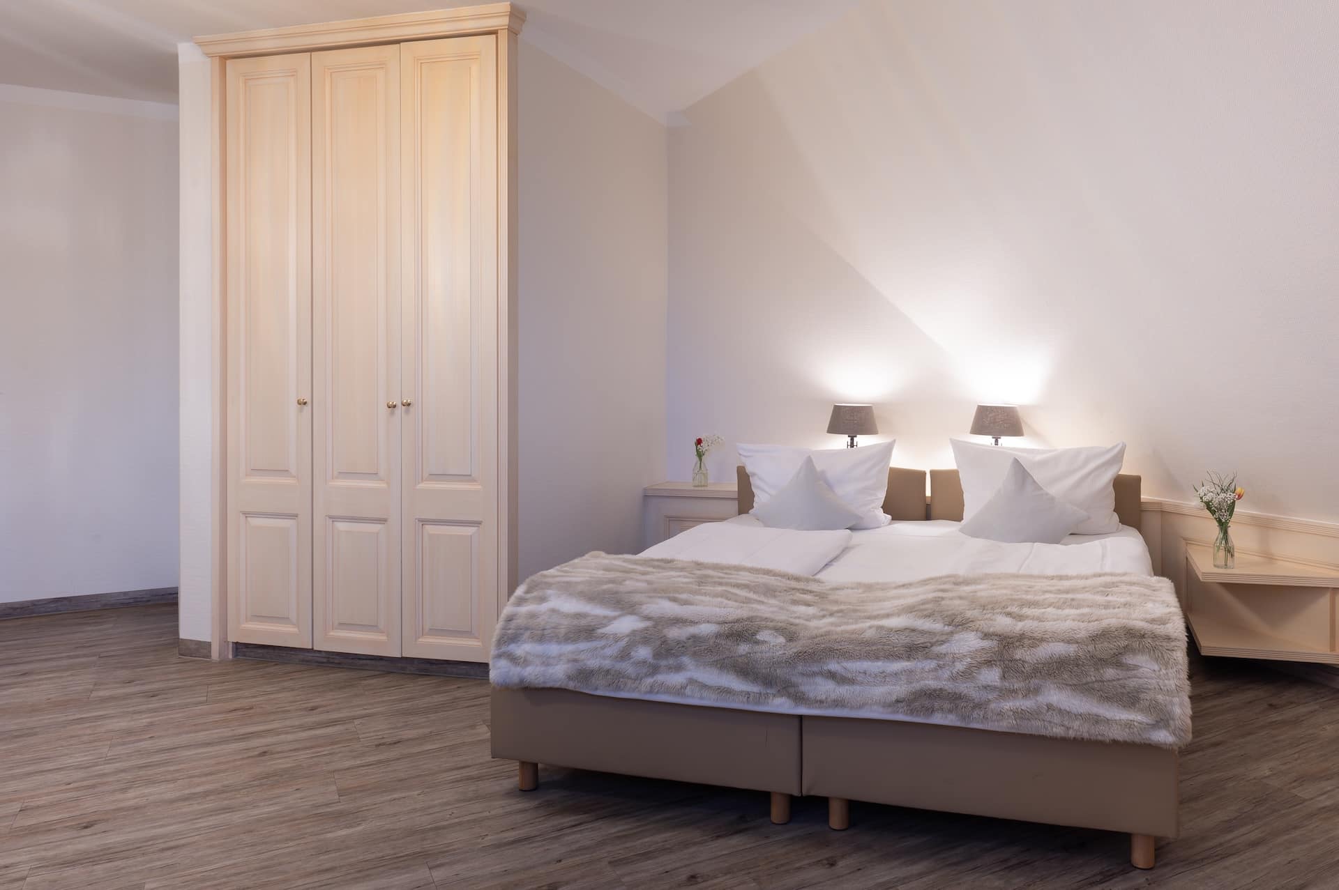 Ein gemütliches Schlafzimmer mit einem großen Bett, das mit einer Pelzdecke bedeckt ist, zwei weißen Kissen, einem hölzernen Kleiderschrank, Nachttischen mit Lampen und Blumen in Vasen. Der Boden ist aus Holz und die Wände sind in einem sanften Beigeton gehalten.