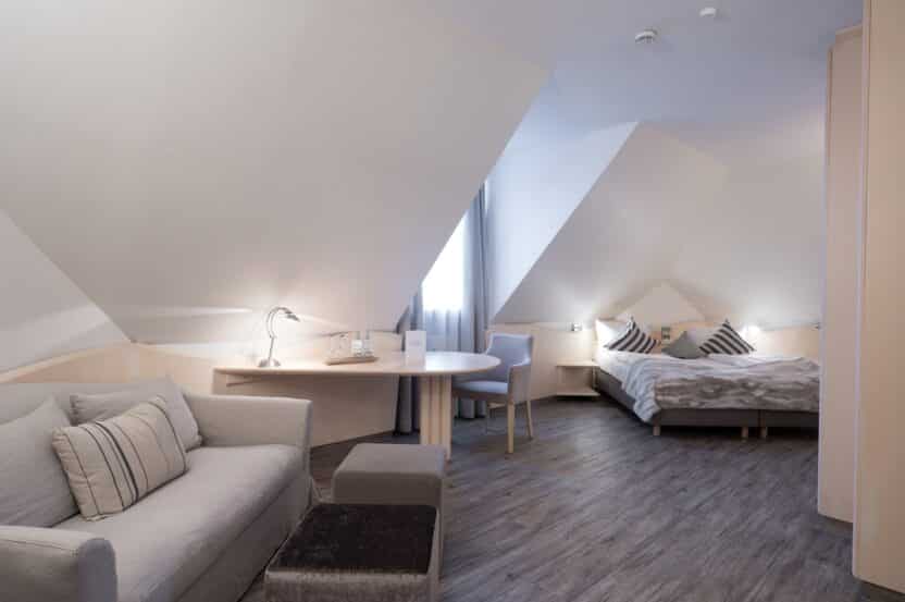 Ein modernes Hotelzimmer mit schrägen Decken, einem Doppelbett mit gestreiften Kissen, einem hellgrauen Sofa mit Kissen, einem kleinen runden Tisch, zwei Stühlen und einem Fenster mit durchsichtigen Vorhängen. Die Einrichtung ist neutral und minimalistisch.