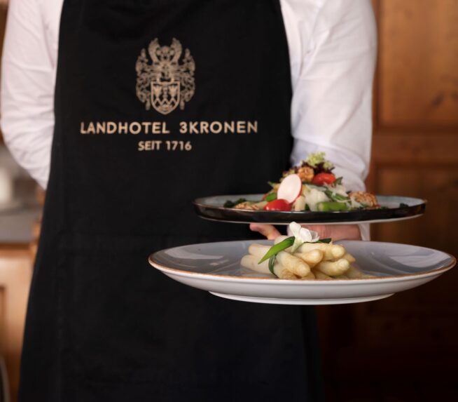 Ein Kellner mit einer schwarzen Schürze trägt zwei Teller mit leckeren Spargelgerichten und fränkischen Spezialitäten