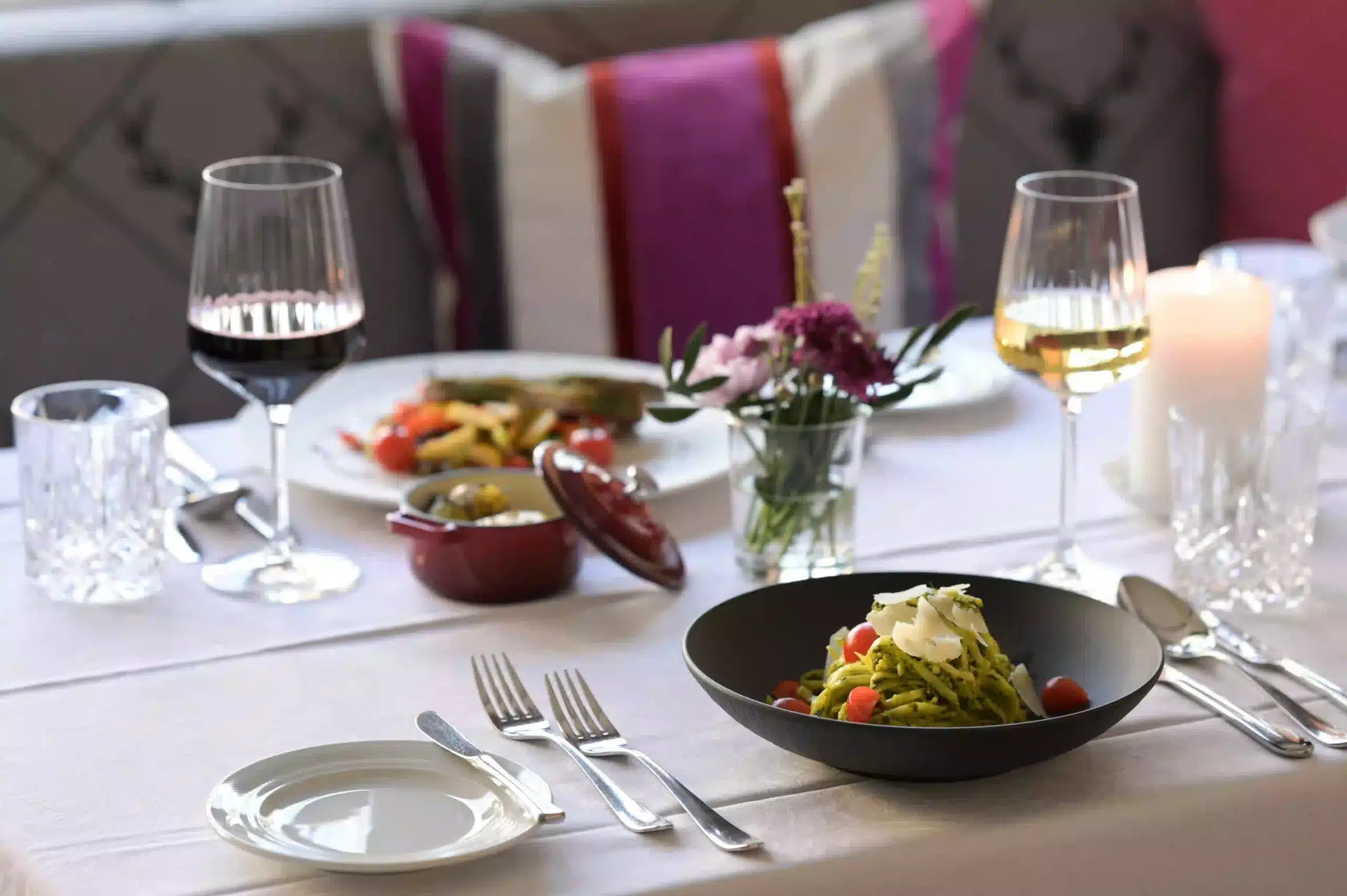 Elegant gedeckter Esstisch mit Tellern mit Speisen, mit Rot- und Weißwein gefüllten Weingläsern, einer kleinen Blumenvase, einer brennenden Kerze und Besteck, ordentlich auf einem weißen Tischtuch angeordnet.