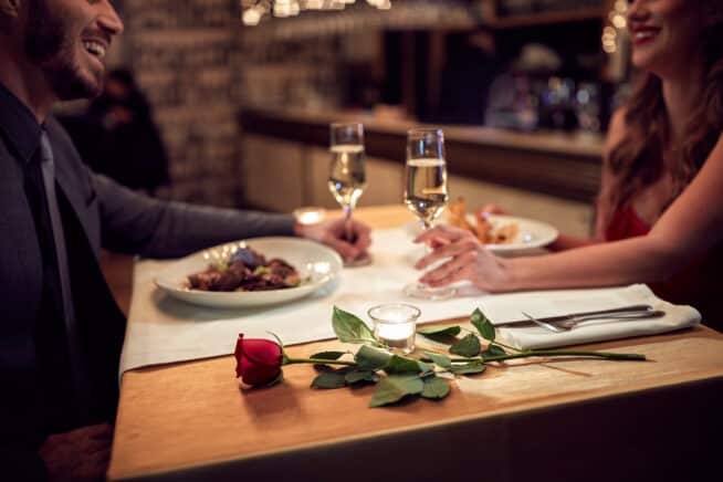 Romantisches Dinner mit zwei Personen, welche sich mit zwei Gläsern Sekt zuprosten und einer roten Rose auf dem Tisch.