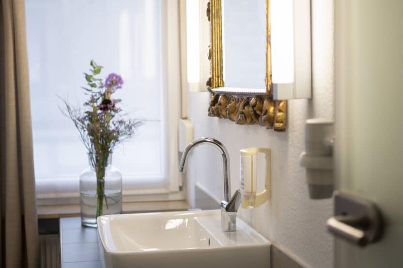 Zimmer 101 Bad: „Helles Badezimmer mit Waschbecken, goldgerahmtem Spiegel und einer Vase mit frischen Blumen im Landhotel 3Kronen.“