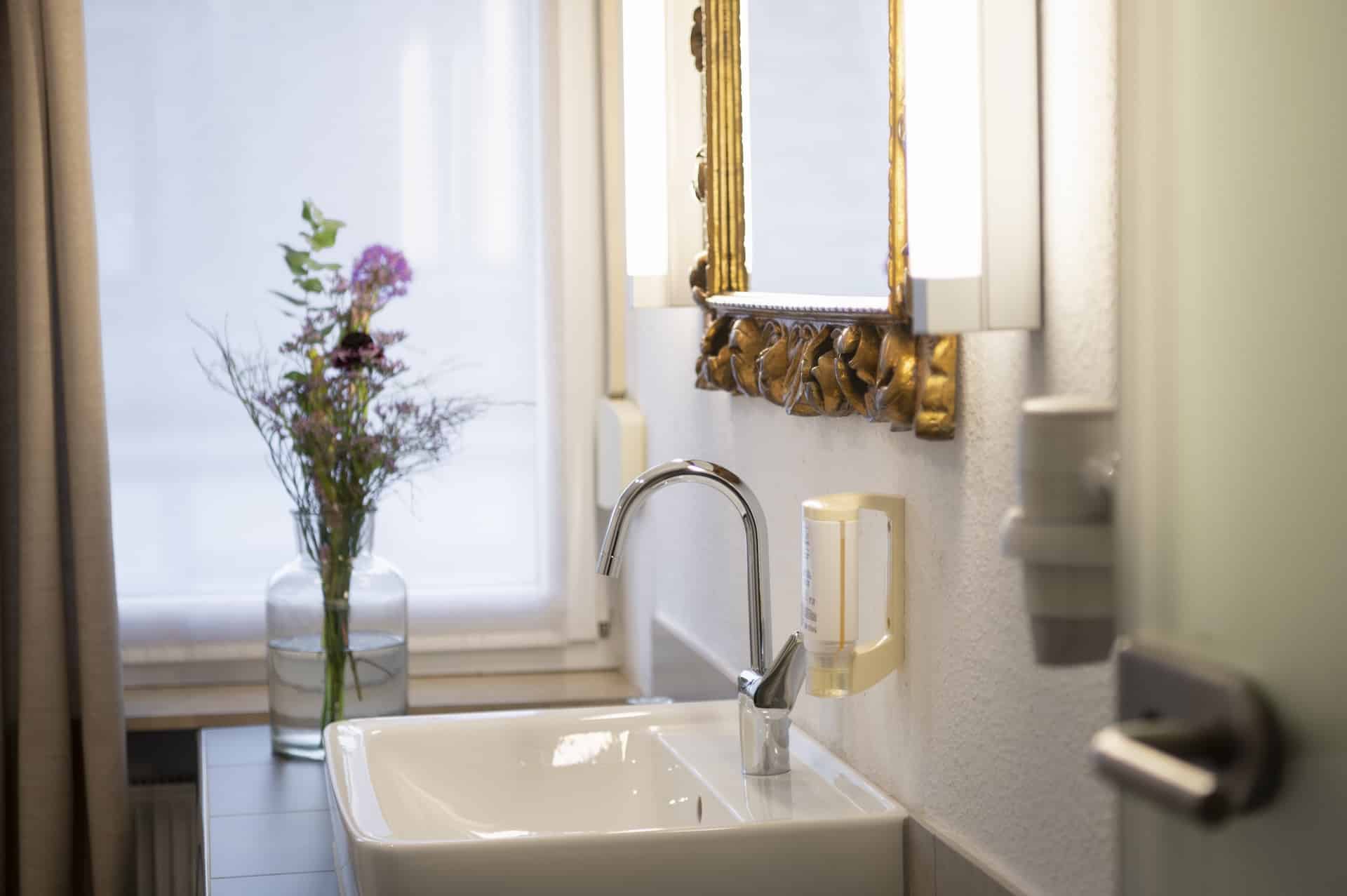 Zimmer 101 Bad: „Helles Badezimmer mit Waschbecken, goldgerahmtem Spiegel und einer Vase mit frischen Blumen im Landhotel 3Kronen.“