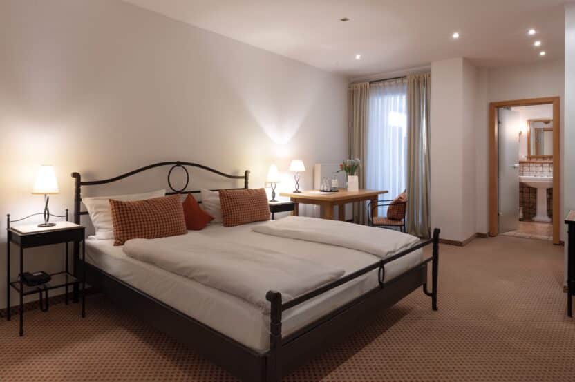 Ein gemütliches Hotelzimmer mit einem Doppelbett, zwei Nachttischen mit Lampen, einem kleinen Schreibtisch mit Stuhl und einem Fenster mit durchsichtigen Vorhängen. Das Zimmer hat eine warme Beleuchtung und ein angrenzendes Badezimmer, das durch eine offene Tür sichtbar ist.